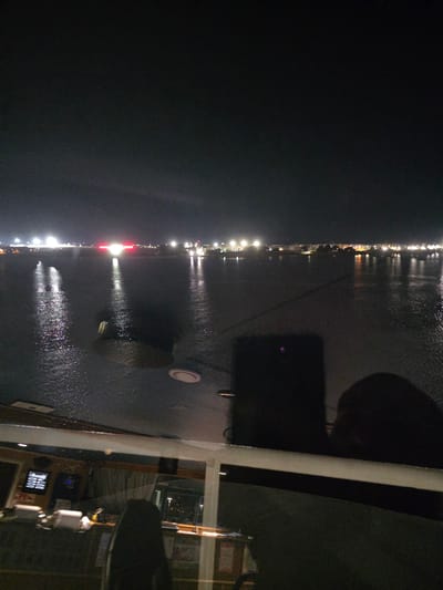This nighttime image captures a view from the bridge of a vessel docked in Brindisi, Italy. The dark expanse of the Adriatic Sea dominates the foreground, reflecting the scattered lights of the distant coastline. The shore is illuminated by numerous artificial lights, including what appears to be a bright, possibly red, marker or beacon in the distance. The lights suggest an urban or industrial area.
On the bridge itself, instruments and control panels are visible, indicating the operational nature of the setting. A black, reflective surface, possibly a screen or window, obstructs a clear view of the environment outside the bridge. The overall scene is one of quiet observation from a sheltered vantage point, overlooking the waters and illuminated city at night.