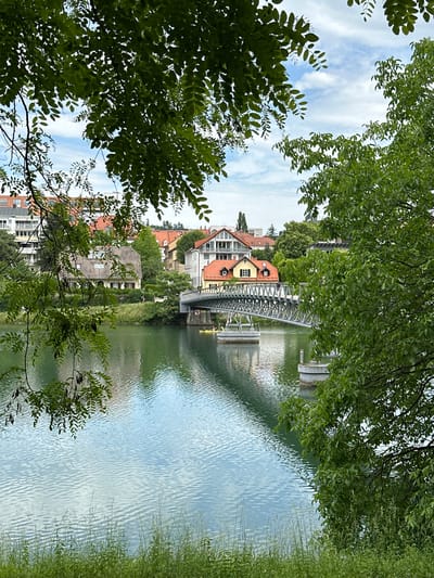 The image depicts a view of the Drava River in Maribor, Slovenia, from a partially obscured vantage point through tree foliage. A steel footbridge spans the river, connecting the banks. The river's surface is rippled, reflecting the sky and surrounding buildings. Residential buildings with orange-tiled roofs line the riverbank. Green vegetation is prominent along the river's edge. Two people are standing on the bridge. The sky is visible through gaps in the trees, displaying a mix of blue and white clouds.