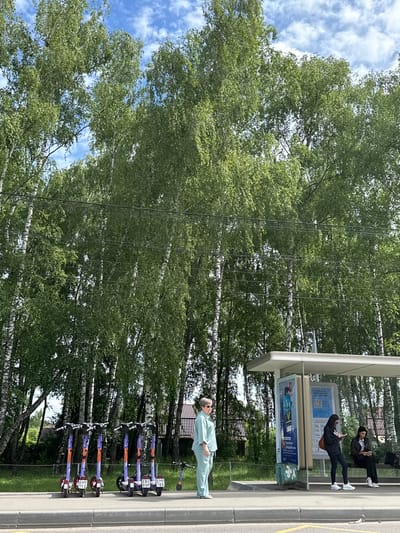 In the city of Berezki SNT (Znamya Oktyabrya), Russia, a bus stop features a modern shelter with advertisements. A woman in light blue loungewear stands to the left of the shelter, looking to the left. To the right of the shelter, two young women are seated on a bench. One is wearing a black hoodie and dark pants, reading a book, while the other is in dark clothing, looking at her phone. In front of the bus stop, several electric scooters are parked, with one further down the road. The background is dominated by a dense grove of birch trees with white trunks, and the sky above is partly cloudy with a blue hue. The ground is paved with a sidewalk and road.