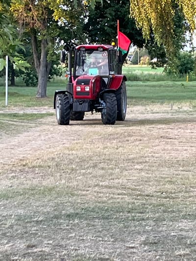 A red tractor with a red flag attached to its cab is positioned in a grassy, outdoor area. The tractor has a driver inside, and its lights are on. The surrounding environment appears to be a rural or park-like setting with trees and dry grass. The image does not provide specific information about the city of Malaryta, Belarus, or any actions or interactions beyond the presence of the tractor and its driver.