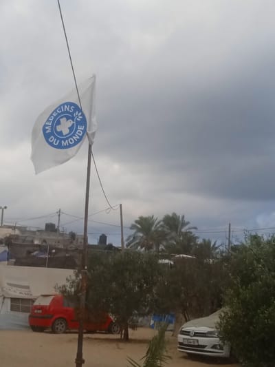The image depicts an outdoor scene in Al-Mawasi, Palestinian Territories, under a heavily overcast sky. A white flag bearing the blue "Médecins du Monde" (Doctors of the World) logo, which features a cross and a dove holding an olive branch, flies on a dark pole in the left-center foreground. Overhead wires are visible, extending from the flag pole and other utility poles. The ground is unpaved, appearing sandy or dusty.
In the midground, several olive-like trees and green foliage are scattered. To the left, a red two-door hatchback car is parked alongside a large white tent, which displays partially visible text, including "AID," suggesting a humanitarian aid structure. To the right, a white Volkswagen sedan is partially obscured by foliage; its Palestinian license plate, "B-1544-04," is discernible.
The background reveals a cluster of low-rise buildings or makeshift structures, many equipped with black water tanks on their roofs. Numerous utility poles and intersecting overhead electrical wires are visible. Behind these structures, several palm trees are present. No people are visible in the scene.
