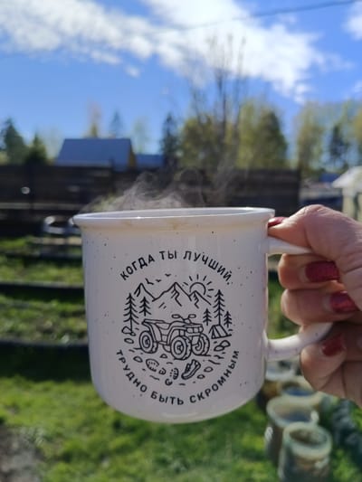 A hand with red nail polish holds a white speckled mug with a black drawing of a mountain scene, an ATV, and the Russian text "когда ты лучший, трудно быть скромным" (when you're the best, it's hard to be humble). Steam rises from the mug. The background shows a blurred outdoor scene with green grass, trees, a wooden fence, and a blue sky with clouds. The location appears to be a residential area, possibly in the city of Вичелово, Russia. Several containers are in the foreground, out of focus.