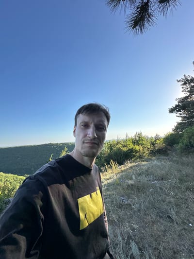 A man stands for a selfie outdoors with a lush green valley and a clear blue sky in the background. The man is wearing a black t-shirt with a large yellow graphic on the front. He is looking directly at the camera with a neutral expression. The sun is shining brightly, casting a warm glow on the landscape. The foreground is dominated by dry grass and some scattered rocks, suggesting a natural, possibly mountainous, terrain. The location is Pryvorottia Druhe, Ukraine.