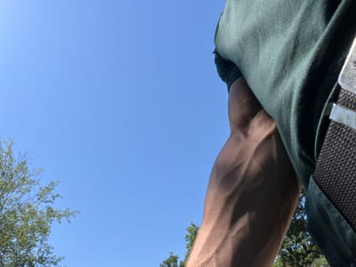 The image shows a close-up, low-angle view of a person's arm and shoulder against a clear blue sky. The person is wearing a dark green garment, possibly a shirt or jacket, which is textured and shows creasing. The arm is muscular and tanned, with prominent veins visible. In the background, parts of trees with green foliage are visible, suggesting an outdoor setting. The sky is a uniform, bright blue with no clouds. The location is identified as Kosti, Bulgaria, implying this scene takes place within that municipality. There are no other individuals or discernible objects that provide further context about the specific activities or interactions occurring. The lighting suggests it is daytime, likely sunny.
