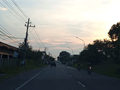 The image depicts a road in Gampongrayadagang, Indonesia, at what appears to be either dawn or dusk, given the soft light and hazy sky. The road is asphalt and has a white dividing line. Several vehicles are visible: a dark-colored SUV is driving in the distance, and at least two motorcycles are also on the road, one further ahead and one to the right. The motorcycle on the right is closer to the camera and its headlight is illuminated. On the left side of the road, there is a building with a fence and overgrown vegetation. Utility poles with numerous wires are prominent along the left side of the road. Trees line the right side of the road, some of which are silhouetted against the bright sky. The sky is a pale orange and white, with scattered clouds.