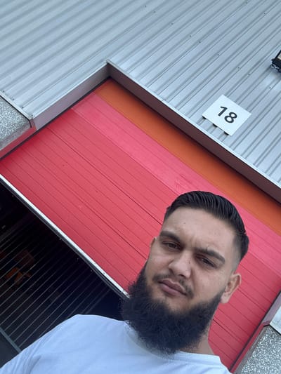 A man with a beard and short dark hair, wearing a white t-shirt, is taking a selfie in front of a large red garage door that is partially open, revealing a dark interior with horizontal shelving units. Above the red door, there is an orange section and then a corrugated gray metal roof. A white square sign with the number "18" in black is affixed to the gray roof. The scene appears to be an outdoor industrial or storage area. The location is identified as Boom, Belgium.