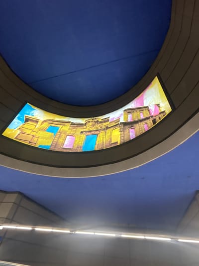 The image shows a curved digital display mounted on a ceiling or overhead structure. The display shows a colorful, stylized depiction of a building with yellow walls and windows of various colors, including blue and pink. The building appears to be rendered in a somewhat abstract or artistic style, with exaggerated perspective and vibrant hues. The background of the display is a bright yellow, and the sky above the building is depicted in shades of blue and white, suggesting a daytime scene.

The display is set against a deep blue background, which appears to be a wall or ceiling surface. Above the blue surface and to the right of the curved display, a series of white fluorescent lights are visible, arranged in a line along a dark ceiling. These lights suggest an indoor environment, possibly a public space like a subway station, airport, or large commercial building. The overall lighting in the image is bright, indicating it is likely daytime. There are no people visible in the image. The scene is static, showing a decorative display in a modern architectural setting.