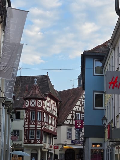 This image depicts a street scene in Ansbach, Germany, likely in the historic city center. In the foreground and left, a tall, light gray banner with dark lettering hangs vertically, displaying the words "HIMMEL MODEHAUS ANSBACH." To the right, several buildings are visible. One building features traditional half-timbered architecture with white plaster and dark wood beams, topped with a red-tiled roof. Adjacent to this, a blue building with white-framed windows stands out. Further to the right, a building with a light-colored facade has a prominent red "H&M" logo sign. A street lamp is mounted on the side of the blue building. The sky above is blue with scattered white clouds. The scene appears to be from a shopping district, with commercial signage visible on some of the buildings.