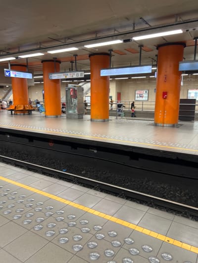 This image depicts the platform of the Horta metro station in Saint-Gilles - Sint-Gillis, Brussels, Belgium. The scene is an underground environment, characterized by a light grey tiled platform with a yellow tactile warning strip running parallel to the metro tracks. Behind this strip, a series of silver, circular tactile indicators are embedded in the tiles.
The platform is supported by multiple large, cylindrical columns painted a vibrant orange, extending from the platform level to the light grey concrete ceiling. The ceiling features numerous rectangular fluorescent light fixtures. Hanging from the ceiling structure are several illuminated signs; one on the left indicates "Horta" and another destination with a blue arrow, while a central sign displays transit-related symbols and text. Another illuminated sign further right shows what appears to be scrolling text.
Along the platform, several orange, circular benches are present around the base of some columns, though no one is actively seated on the fully visible bench. A metallic grey emergency cabinet with a red panel is situated between two columns. A small red box, possibly an emergency stop button, is affixed to the rightmost orange column.
Beyond the platform, the metro tracks are visible, consisting of two metal rails laid on dark ballast. In the background, a light-colored tiled wall with posters or advertisements is visible above a short railing. An upward-sloping structure with handrails, indicative of a stairwell or escalator, leads to an upper level.
Several individuals are present:
* On the left side of the platform, near the orange benches, indistinct figures are visible, including one person in blue.
* In the mid-ground, near the ascending stairwell, a person in dark clothing walks towards the right, slightly behind and to the right of a smaller individual (a child) in lighter-colored clothing (pink/red top).
* Additional indistinct figures are visible further in the background near the top of the stairwell.
No direct interactions between individuals are discernible; most appear to be moving or waiting.