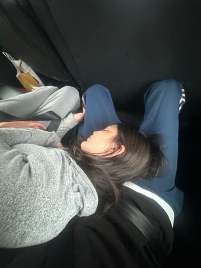 The image captures an interior vehicle scene, reportedly in Bondy, France, from a high-angle perspective. Three individuals are partially visible within what appears to be the rear passenger compartment of a car. A young individual with long, dark hair, wearing a grey marled hooded sweatshirt and grey sweatpants, lies horizontally in the foreground. Their eyes are closed, indicating sleep or rest, with their head positioned on the lap of a second individual. This second individual, largely obscured, is wearing dark blue track pants featuring white stripes along the outer seams, and a black seatbelt is fastened across their lap. A third individual is partially visible on the far left, wearing grey sweatpants, with a black seatbelt also secured across their lap and a hand resting on their thigh. A light-colored paper bag with brown handles is situated near this third individual. The vehicle's interior is predominantly dark, providing minimal environmental detail beyond the occupants and their immediate surroundings.