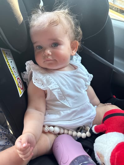 A close-up, eye-level shot captures a baby girl lying on her side in a car seat. Her blonde hair is slightly messy, and her large blue eyes are looking directly at the camera. She has a fair complexion with rosy cheeks and a small smile. The baby is wearing a white eyelet lace top with ruffled sleeves.

In the background, a plush Santa Claus toy is visible, partially obscuring the baby's shoulder. A pink sippy cup or bottle is also present, positioned near the baby's leg. A string of large, light-colored wooden beads is draped over the baby's chest, attached to something out of frame.

The car seat is black, and a yellow and black warning sticker is visible on the seat, reading "AIRBAG" with a graphic indicating a child should not be in the front seat. The lighting suggests it is daytime, with sunlight casting shadows on the car seat. The overall scene conveys a sense of a family outing or travel, with the baby appearing content and alert. The location context of Giessen, Germany, implies the setting is likely within a car in that region.