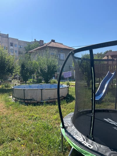 The image depicts a backyard scene in Belene, Bulgaria, featuring a trampoline and a round above-ground swimming pool. The trampoline, branded "ZIPRO," is in the foreground and has a safety net enclosure. Adjacent to the trampoline, to the right, is a blue slide, likely part of a children's play structure. In the middle ground, a circular swimming pool with a blue patterned interior is visible. The yard is covered in green grass and some patchy dry areas. Behind the yard, there are residential buildings, including a multi-story apartment building and a house with a tiled roof. Trees and bushes are scattered throughout the scene. Laundry is hanging on a clothesline in front of one of the buildings. The sky is clear and blue, indicating a sunny day.