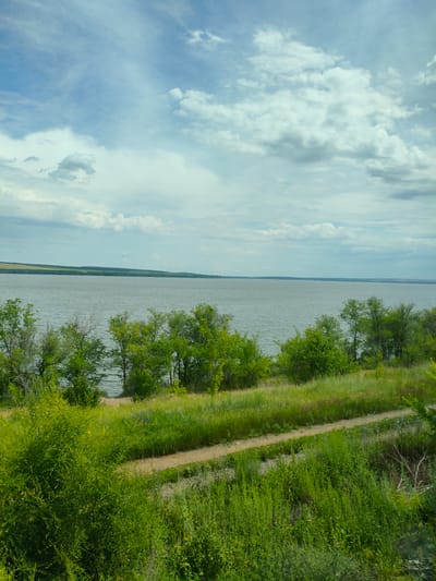 This image depicts a wide, calm body of water, likely a river or lake, under a partly cloudy sky. The water is a muted gray-blue, reflecting the overcast conditions. Along the near bank, a line of lush green trees and vegetation slopes down towards the water's edge. In the foreground, a dirt path winds through tall green grass and bushes. The landscape appears to be natural and undeveloped. The lighting suggests it is daytime, likely late afternoon or early evening given the soft, diffused light. The scene evokes a sense of peace and tranquility. The location is identified as Buzuluk, Russia. There is no visible text in the image.