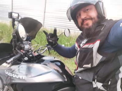 A man wearing a motorcycle helmet and a vest is sitting on a motorcycle. The man is holding up two fingers in a peace sign. The motorcycle is a dark color and has a windshield and mirrors. There is a camera mounted on the motorcycle. The background is a metal fence and some grass. The man appears to be in Semypolky, Ukraine.