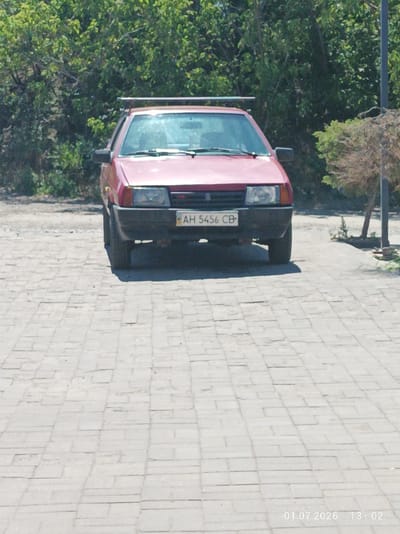 A red car is parked on a paved surface. The car has a license plate that reads "AH 5456 CB". In the background, there are green trees and bushes. The paved surface appears to be cobblestone or brick. The scene is outdoors and the lighting suggests it is daytime. The date and time stamp at the bottom right corner indicates July 1, 2026, at 13:02. The car is oriented facing forward.