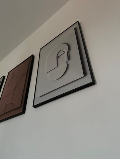 The image shows a close-up of two framed pieces of abstract art hanging on a white wall. The art on the right is a grey, layered geometric design that resembles a stylized face or head. The art on the left is a brown, layered abstract design with curved lines. Both pieces are framed in black. The image is taken from a low angle, looking up at the wall and the artwork. The scene appears to be indoors, likely in a home or gallery. There are no people or animals visible in the image. The wall has a slightly textured finish. No specific location in Darlington, United Kingdom is identifiable from the image.