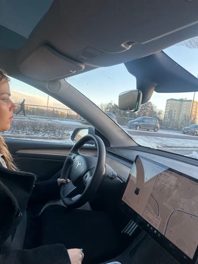 The image displays the interior of a Tesla Model 3 or Y, viewed from the front passenger seat, looking forward and slightly left. A female driver with brown hair is visible in profile on the left, wearing a dark coat and a light-colored top, with her left hand on the black multi-function steering wheel. Her right hand, wearing a ring with a green stone, is visible in the lower right.

The car's large central touchscreen displays a navigation map indicating the location in Solna, Sweden, with "Råsundavägen" and "Solna Centrum" clearly visible as street and area names. The map shows a route with a destination 900 meters away, an estimated arrival time of 19:00, and the current vehicle speed as 38. A Spotify icon is present on the screen.

Outside the vehicle, the ground and roadside areas are covered in snow and ice, indicating winter conditions under a clear blue sky. In the mid-ground to the left, a pedestrian in dark clothing is walking left on an elevated bridge or overpass. Several other vehicles, including a silver hatchback, are visible on the road in the background. A multi-story residential building with a light-colored facade stands in the right background. Bare trees and shrubs line the road. The windshield has some visible ice or dirt along its lower edge. A panoramic glass roof is visible above the passenger sun visor.
