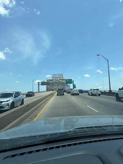 A frontal view from inside a vehicle shows traffic on a multi-lane bridge. The sky is bright blue with scattered white clouds. On the left side of the image, a silver SUV is driving in the adjacent lane. The bridge has a concrete barrier separating the lanes, with a yellow line marking the edge of the roadway. In the distance, the bridge's metal structure is visible, along with overhead signs. One sign indicates "EXIT 159A TO LSU," and another lists "Nicholson Dr HIGHLAND RD WEST." Several other vehicles, including cars and trucks, are on the bridge. A street lamp is visible on the right side of the bridge. The foreground shows the dashboard and windshield of the vehicle. The scene appears to be within the city of Port Allen, Louisiana, United States of America.