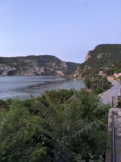 The image shows a scenic view of the Danube River at dusk, with the Golubac Fortress visible in the background. The river is wide and calm, with a few small boats dotting the surface. The surrounding landscape is dominated by lush green hills and rocky cliffs, characteristic of the Iron Gate gorge. A winding road runs along the riverbank, with a few houses nestled among the trees. The sky is a soft gradient of blue and purple, indicating the transition from day to night. The overall atmosphere is peaceful and serene. The scene is located in the Golubac Municipality of Serbia.
