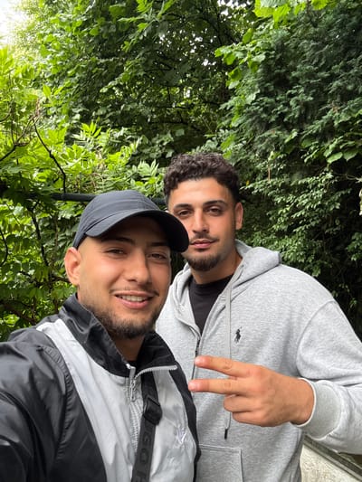 Two young men are pictured outdoors in what appears to be a natural setting with abundant green foliage and trees, possibly in Liestal, Switzerland. The image is a close-up, likely a selfie, taken from a slightly upward angle, capturing the faces and upper torsos of both individuals.
In the foreground, a man with a dark baseball cap is smiling broadly, revealing his teeth. He has dark facial hair and brown eyes. He is wearing a black and white jacket with a zipper, and a black strap or bag is visible over his shoulder with the partial text "HUGO" (likely Hugo Boss) printed on it.
Behind him, slightly to the right, another man with curly dark hair, a beard, and brown eyes is also looking towards the camera. He is wearing a light grey hooded sweatshirt with a small, dark polo player logo embroidered on the left chest area, resembling a Ralph Lauren logo. His right hand is raised, making a peace sign (V-sign) with his index and middle fingers.
The background is filled with various types of green leaves and branches, indicating a lush, natural environment. The lighting suggests it is daytime. Both men appear happy and relaxed, with smiles on their faces.