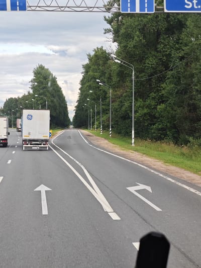 The image shows a two-lane road in Polustovo, Russia, with a dense green forest on the right side and trees lining the left side of the road. Traffic lights are visible in the distance, and streetlights are positioned along the right side of the road. The road has white lane markings, including arrows indicating a straight path and a turn to the left. Several trucks are on the road; one truck in the foreground has a "GE" logo and a license plate. The sky is overcast with some patches of blue. A blurry black object, possibly a rearview mirror, is visible in the lower right corner of the image.