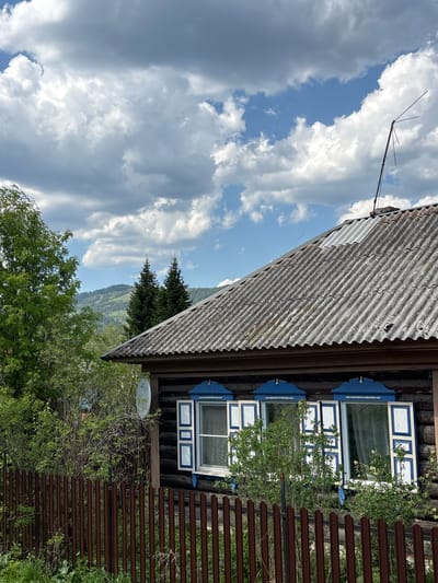 The image captures a rustic scene likely in a rural area, possibly Овсянка, Russia, as indicated in the prompt. The scene includes elements of nature and a wooden structure with distinct architectural features.
A corrugated metal roof dominates the top portion of the image. Underneath, a dark wooden wall has windows with white frames trimmed with blue accents. Vegetation, including climbing plants, partially covers the windows and walls.
To the side, a wooden fence made of horizontal slats stretches from the side. The background features rolling hills and trees under a sky filled with clouds. An antenna is visible on the roof, suggesting the presence of technology amidst the natural setting.
