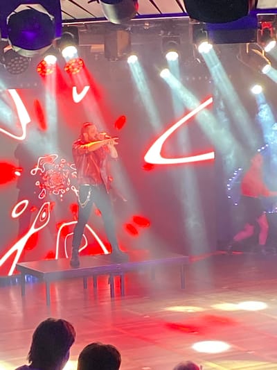 This image depicts a live performance taking place on a stage. A male performer, dressed in a red leather jacket, black pants, and a silver chain belt, is center stage, holding a microphone and singing. He is standing on a raised platform. Behind him, a large screen displays abstract red and white graphics. Stage lighting, predominantly red and white, illuminates the scene, creating a smoky atmosphere. To the right of the main performer, a second performer, wearing a red top and black skirt, is visible, possibly engaged in a dance or prop manipulation. The stage floor is illuminated by circular light projections. In the foreground, the backs of two audience members are visible, indicating the presence of a crowd. The overall environment suggests an indoor concert or performance venue. There is no information in the image or provided crops to identify the specific location in the city of Hitis, Finland.