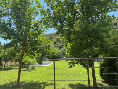 A sunny outdoor scene features a lush green lawn, two trees with vibrant green foliage, and a clear blue sky. In the foreground, a modern metal railing with horizontal bars partially obstructs the view of the lawn and trees. Beyond the lawn, a small white structure, possibly a pool or fountain, is visible, along with more trees and a sloping hillside covered in dense vegetation. The environment suggests a well-maintained outdoor space in a rural or suburban setting.