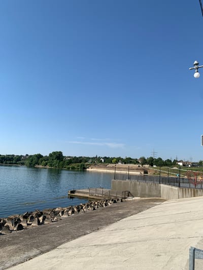 The image shows a bright, clear day at a waterfront with a paved embankment. The water is calm, with some vegetation floating on the surface. Along the edge of the water are triangular concrete anti-tank obstacles. Further up the embankment, there is a concrete wall with a metal railing and several lamp posts. In the background, across the water, is a tree-lined shore with houses and buildings. Power lines and poles are also visible in the distance. The sky is a solid, cloudless blue. The scene is a public space, likely a park or promenade, designed for recreation and to manage the shoreline.