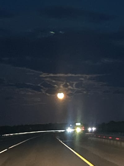 The image depicts a highway at night, likely in or near Tyukalinsk, Russia, illuminated by the headlights of passing vehicles and the bright glow of a partial solar eclipse. Dark clouds obscure most of the sky, but a segment of the sun is visible, appearing as a luminous, crescent-shaped object. The highway stretches into the distance, with lane markers clearly visible. Multiple cars are driving in both directions, their headlights creating streaks of light. A few trees and structures can be seen along the roadside, silhouetted against the dim light. The overall atmosphere is dark and somewhat mysterious, with the celestial event adding an unusual element to the nocturnal scene.