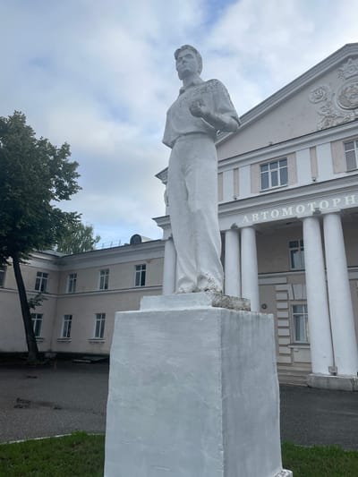 The image shows a white stone statue of a young man in work clothes standing on a white pedestal in front of a building in Zavolzhye, Russia. The statue is in the foreground, occupying the right half of the frame. The man is depicted in a dynamic pose, with his right hand raised to his chest and his left hand holding what appears to be a small object. He wears a collared shirt and loose-fitting trousers. The building behind the statue is a multi-story structure with a neoclassical facade, featuring white columns and large windows. The words "АВТОМОТОРН" are visible in large letters above the columns. To the left of the statue, a large tree with dark green foliage partially obscures the building. The ground in front of the statue is paved with asphalt and grass. The sky is overcast with clouds.