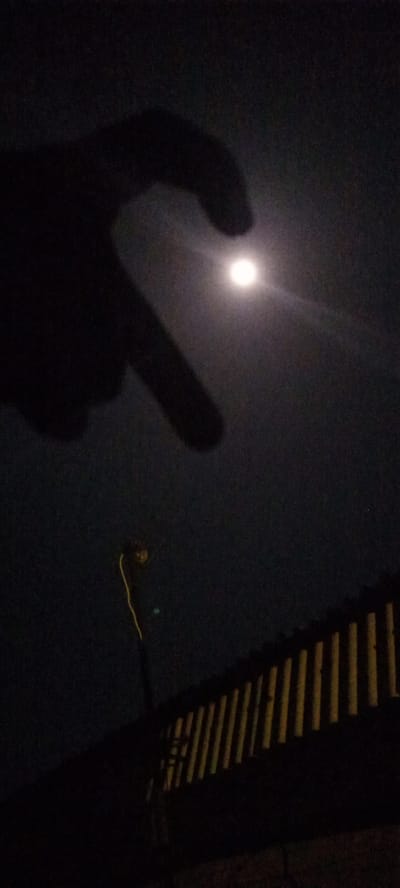 The image captures a nighttime scene in Ilogbo Eremi, Nigeria, dominated by a large, dark silhouette of a hand in the foreground. The hand is positioned to form a partial heart shape, with the fingers curving towards the center of the frame. Through the opening created by the hand, a bright, circular full moon is visible, emitting a soft glow and appearing as the focal point.
In the lower half of the image, to the left, a slender utility pole or street light is dimly visible, with a yellow-orange wire extending upwards from it. To the right, a fence or railing with vertical, evenly spaced slats is illuminated from behind or from a low angle, casting distinct, warm-toned stripes across the dark background. The sky is dark and clear, suggesting good weather conditions, allowing the moon's brightness to stand out. The overall mood conveyed is one of contemplation or appreciation for the night sky, emphasized by the heart-shaped gesture framing the moon. There is no visible text in the image.