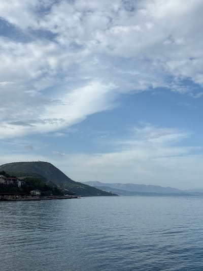 The image shows a landscape of a coastline. On the right is water, reflecting light. To the left of the water is a green mountain with buildings at its base, possibly a town or village. Behind the initial mountain, a range of mountains fades into the background. The sky is a blend of blue and white, with clouds scattered throughout. The location is not apparent from the image and cannot be ascertained.