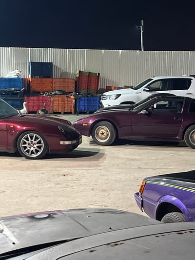 The image shows an outdoor parking or storage area at night, rotated 90 degrees counter-clockwise. The location is Kfar Qasim, Israel.

In the center-left, a white Toyota Hilux pickup truck is parked, with "HILUX" visible on its side. Next to it, a deep purple sports car, possibly a Nissan 300ZX, is visible with gold mesh-style wheels and a pop-up headlight partially open.

In the immediate foreground, a maroon or reddish car, also potentially a Nissan 300ZX but a different generation, is parked with multi-spoke silver wheels. A small, grey and white cat is seen sitting on the driver's side window frame of this car, looking to the left.

On the far right, the front corner of a bright purple car with a distinctive lime green stripe is visible, possibly a classic American muscle car. Another grey-colored vehicle is partially visible further right.

The background features stacks of colorful plastic crates in orange, blue, and red, along with some wooden pallets. A dark sky and a corrugated metal wall or building are also visible. The ground is a light-colored paved surface, likely concrete. No people are visible.