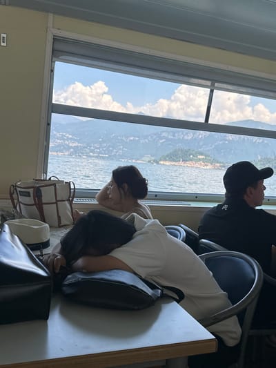 Two people are resting on a ferry in Tremezzina, Italy. The person in the foreground, a woman, is leaning on her arm, with her head resting on a black purse. She is wearing a white top. Beside her is another person, also a woman, leaning forward with her head down and out of frame. In the background, a man wearing a black baseball cap and black sweatshirt is seated and looking out the window. The ferry is moving, with the water of a lake visible outside the large window. Mountains and a small town can be seen across the water. The sky is blue with fluffy white clouds, indicating it is daytime and the weather is fair. The scene suggests a moment of relaxation and travel, possibly during a vacation. There is no visible text in the image.