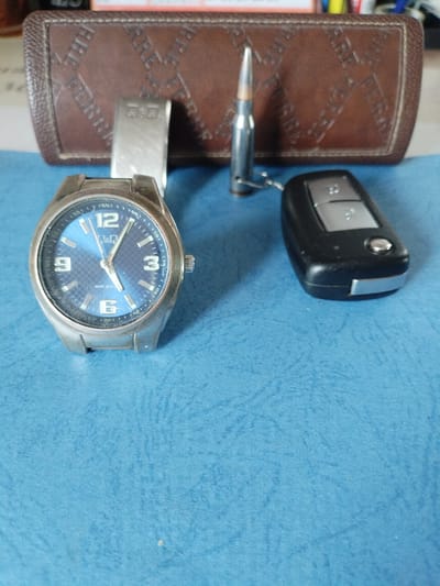 A Q&Q brand wristwatch with a blue analog dial and a metal band lies on a blue textured surface, with its face angled towards the viewer. To the right of the watch, a black car key fob with silver accents is placed. Behind the watch and key fob, and slightly out of focus, is a brown leather-like case with embossed lettering and a single metallic bullet standing upright with a keychain attached. The background is a blurred brown case with embossed lettering. This scene is located in the Вароніцкі сельскі Савет, Belarus.
