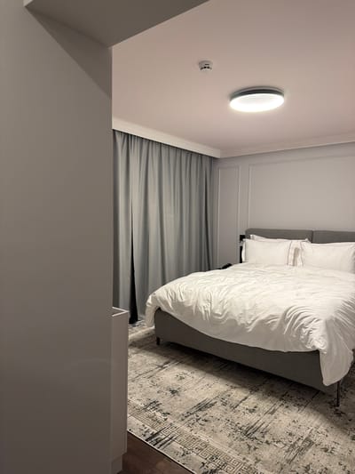 This media file captures a modern bedroom interior, presented in a portrait orientation that appears to be rotated 90 degrees clockwise from its original landscape perspective.
The central subject is a bed, featuring a gray upholstered headboard and a crisp white duvet with matching pillows. A textured rug with an abstract and geometric pattern in shades of gray and off-white is visible on the floor beside the bed.
On the wall above the bed, a circular, illuminated wall light fixture emits a bright, white glow, suggesting the room is lit by artificial light, possibly indicating evening or night. Directly below the light, a white smoke detector is mounted on the wall. The walls are painted a light neutral color and feature simple decorative molding or paneling.
To the side of the bed, dark gray curtains are drawn, covering a window. The overall aesthetic is minimalist and contemporary, with a dominant monochromatic color scheme of grays and whites.
No people are present in the image, and no specific activity is taking place. There is no visible text.