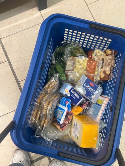 A high-angle shot captures the interior of a blue plastic shopping basket, filled with various groceries, positioned on a light-colored, speckled tiled floor with gray grout lines. The scene is located within a supermarket in Heemskerk, Netherlands.

Inside the basket, visible items include: a clear plastic bag of green leafy vegetables (likely spinach), two clear plastic bags containing gnocchi or similar potato dumplings, a stack of packaged cookies or biscuits, two containers of "Goldessa" branded cream cheese or spread (one 200g, one 300g), a small blue and white carton, a vacuum-sealed block of yellow cheese, and a package of imitation crab sticks (surimi). Other less identifiable plastic-wrapped items are also present.

The lower portion of a person's legs and feet, wearing white sneakers and light-colored trousers or jeans, is visible at the bottom of the frame, indicating the person is standing near or holding the basket. A dark handle of the shopping basket is visible on the left side. The lighting appears to be ambient indoor illumination.