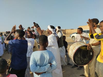 A group of people, mainly men and boys, are gathered outdoors in a dusty, open area. Some are clapping their hands, while others are playing a large drum. Many wear traditional clothing, including white robes and head coverings. In the background, there is a white vehicle and a building, suggesting a village setting. The sky is clear and light blue, indicating it is daytime. The atmosphere suggests a celebration or a gathering with music and communal participation.