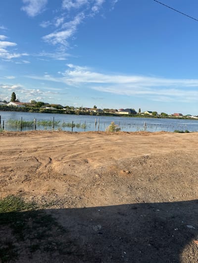 The image depicts a waterfront scene in the village of Құрманғазы ауылдық округі, Kazakhstan. In the foreground, there is a strip of dry, sandy ground with tire tracks and some scattered green vegetation on the left. This area appears to be a dirt road or embankment. Beyond this, a body of water, likely a river or lake, stretches across the middle ground. A series of wooden posts are partially submerged in the water, forming a rough line. Some reeds and aquatic plants are visible in the water near the posts. In the background, across the water, is a residential area with several houses, trees, and vegetation lining the shore. The sky above is blue with scattered white clouds, suggesting it is daytime. A power line is visible stretching across the top right corner of the image. There are no people or animals visible in the scene. The overall impression is one of a rural or semi-rural settlement by the water on a clear day.