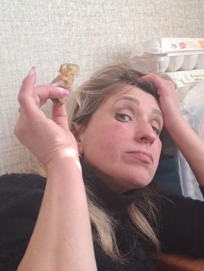 Within a domestic or residential setting in Bolshaya Elhovka, Russia, a single adult female is depicted in a close-up, chest-up shot. The woman, with blonde hair and a fair complexion, wears a dark, possibly black, high-necked garment. Her facial expression is one of apparent exasperation or distress, characterized by slightly parted lips, down-turned mouth corners, and flushed cheeks. Her eyes are directed towards the camera. Her right hand (viewer's left) is elevated, holding a small, irregularly shaped, light-brown, textured object, consistent with a piece of food, between her thumb and index finger. Her fingernails are manicured with a pink and white French tip design. Her left hand (viewer's right) is placed on her head, fingers touching her forehead area. The background features a light-colored, textured wall. To the right, stacked white cardboard egg cartons, bearing Cyrillic inscriptions, are visible.
