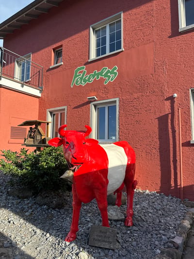 A bright red, stylized cow statue with white patches stands in front of a textured, red building. The cow has a large bell around its neck. A sign at the base of the cow reads "Resi von der Felsenegg". The building has several windows and a green, stylized sign that reads "Felseneck". To the left of the cow, a wooden birdhouse is mounted on a post near a green bush. The ground is covered in gravel and stones. The scene appears to be outdoors, under daylight. The building is located in Stallikon, Switzerland.
