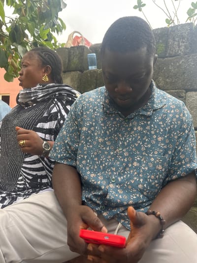 The image shows two individuals, a woman and a man, seated outdoors in Ganyingbo, Nigeria. The woman, on the left, has braided hair and is wearing a black and white patterned scarf and a black and white patterned garment adorned with what appear to be rhinestones. She has on gold leaf-shaped earrings and two gold rings on her left hand, one of which is a double band. A watch with a dark face and metal band is visible on her wrist. She is looking to her left, with her mouth slightly open.
The man, on the right, is wearing a teal collared shirt with a floral pattern in shades of beige and brown. He is looking down at a red smartphone held in his hands. He has a bracelet made of dark, round beads on his right wrist. Both individuals are wearing light-colored pants. They are seated in front of a wall constructed of rough-hewn, grey stones. Vegetation, including green leaves, is visible in the upper left and upper right corners of the frame. The sky appears overcast.