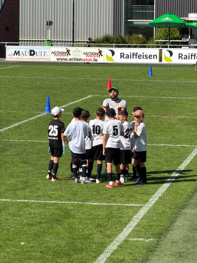 The image shows a soccer team of young players, likely in Fentange, Luxembourg, based on visible advertising signage. They are lined up on a grass soccer field along a white line, with a coach standing with them. The players are wearing white jerseys with black shorts, numbered 25, 10, 5, and 27. The coach wears a grey shirt and a green hat and is giving a thumbs-up gesture. There are orange and blue cones positioned at either end of the team. In the background, there are advertising signs for "MAUDUITZ", "ACTION WEAR", and "Raiffeisen", as well as a "BOFFERDING" umbrella. A building with corrugated siding is visible on the left side of the image. The lighting indicates a sunny day.