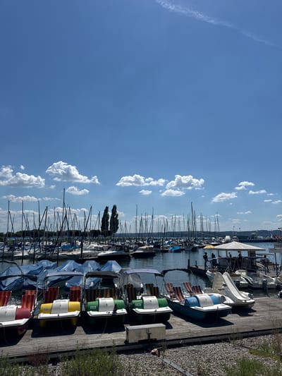 The image shows a marina with several boats docked on a wooden pier. The boats are of various sizes, including sailboats and what appear to be pedal boats or small recreational boats. Some of the boats are covered with tarps, while others are uncovered. The scene is set under a clear blue sky with scattered white clouds. The time of day appears to be daytime, likely late morning or afternoon, given the bright sunlight casting shadows.

In the foreground, there are several colorful pedal boats lined up on the pier, some with numbers visible on them, such as "3", "7", and "5". They have striped seating in red, yellow, and blue. Further back, larger sailboats with tall masts are docked in the water. A large white tent structure, possibly a shelter or part of a building, is visible over some of the boats. In the background, more boats and structures are visible along the waterfront, with trees and a distant hillside.

The overall atmosphere is one of a calm and sunny day at a recreational marina. There are no people prominently featured in the image, though some figures can be seen in the background near the tent structure. The location context indicates Uhldingen-Mühlhofen, Germany, suggesting this is a harbor on a lake or river in that region. The image is oriented vertically.
