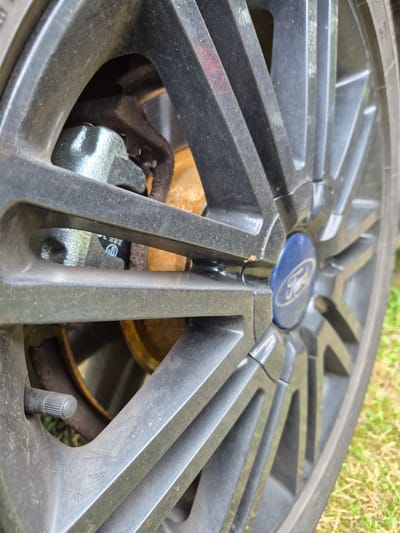 This is a close-up, low-angle shot of a dark gray, five-spoke alloy wheel with a blue Ford center cap. The wheel appears to be dirty and scratched in places. Through the spokes, a rusted brake rotor and a silver-colored brake caliper are visible. There is also a black valve stem with a ribbed cap. The background is out of focus, showing green grass and some dirt. The image was taken outdoors during the day. The location is Masów, Poland. There is no discernible text in the image, other than a small marking on the brake caliper which is too blurry to read.