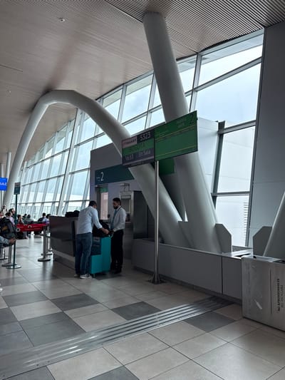 The image captures a modern airport gate area, specifically Gate 2, within Matecaña International Airport (PEI) in Pereira, Risaralda, part of the Area Metropolitana Centro Occidente (AMCO), Colombia. The interior features a high ceiling with prominent white curved structural beams and expansive glass curtain walls, allowing natural light and a view of an overcast sky. The floor is composed of light and dark gray square tiles, with a tactile paving strip in the foreground.
In the mid-ground, a male passenger in a light blue long-sleeve shirt and jeans is positioned at a check-in or boarding counter, placing or adjusting a black bag on a teal-colored baggage sizer/scale marked "Smart MAX.3000." Opposite him, behind the counter, stands a male airline staff member with a dark beard, wearing a white collared shirt, dark trousers, and a light blue tie, observing the passenger's action.
To the right of the counter, two digital display screens are mounted on a pole. The left screen displays flight information: "JetSMART 5323" to "BOGOTA," time "16:53," and status "En Sala" (Boarding). The right screen indicates "PUERTA GATE 2." In the background, multiple individuals are seated on red airport waiting chairs. A stainless steel waste receptacle, labeled "NO APROVECHABLES" and "NO RECYCLABLE," is visible on the far right.