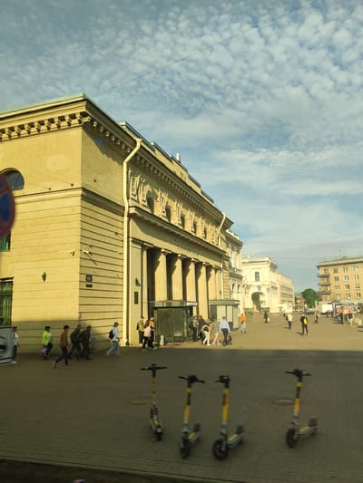 The image captures a street scene in Saint Petersburg, Russia, likely on a sunny day with scattered clouds. In the foreground, three yellow and gray electric scooters are parked on a paved sidewalk. Beyond the scooters, a group of people are walking, some individually and some in small groups.
The main architectural feature is a large, light-yellow building with classical elements, including columns and decorative carvings along its upper facade. A rounded window is visible on the corner of the building. Several people are seen near the entrance of this building, which appears to be a public building, possibly a metro station, given the signage visible.
Further down the street, other buildings are visible, including a multi-story apartment building on the right with a light-colored facade and numerous windows. The street is paved with interlocking bricks, and power lines are strung across the sky. The overall impression is a busy urban environment with a mix of historical and modern architecture.