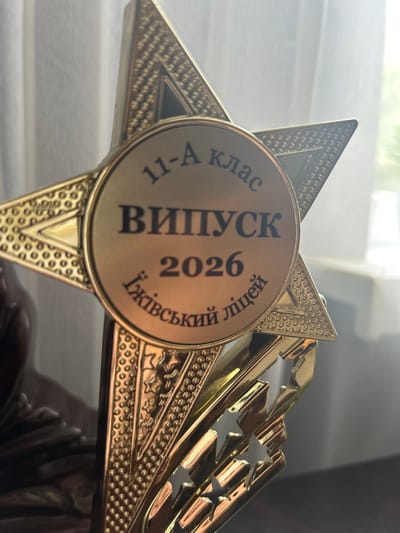 The image is a close-up shot of a gold-colored star-shaped trophy, likely awarded for academic achievement. The trophy features a circular medallion in its center, inscribed with Ukrainian text. The top arc of the medallion reads "11-А клас", indicating it's for the 11th grade, class A. Below this, in larger letters, is "ВИПУСК", meaning "Graduation". Underneath the graduation text is the year "2026". The bottom arc of the medallion bears the inscription "Їжівський ліцей", identifying the institution as the Yizhivtsi Lyceum. The trophy is highly detailed with embossed patterns and star cutouts on its pointed extensions. The background is blurred, showing white curtains and a hint of green foliage, suggesting an indoor setting near a window. The lighting is soft, casting a warm glow on the metallic surface of the trophy. There are no people or animals visible in the frame. The trophy is positioned on a dark, reflective surface, possibly a table or shelf, on which faint reflections of an unknown object can be seen.