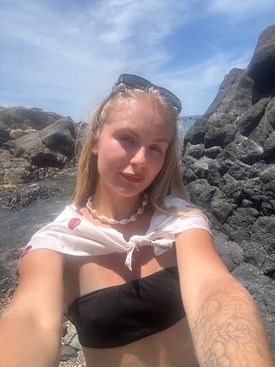 A young woman with blonde hair and a shell necklace is taking a selfie outdoors. She is wearing a white bikini top with a cherry print and a black bikini bottom. Sunglasses are perched on her head. Her arm, which has a tattoo, is visible.
She is standing in a rocky, coastal environment. Large, dark rocks are present behind her and to the side, with the ocean visible in the background. The sky is blue with some clouds. The lighting suggests it is daytime, likely sunny. The overall mood appears relaxed and happy.
The location is identified as Phú Lương, Vietnam. There is no visible text in the image.